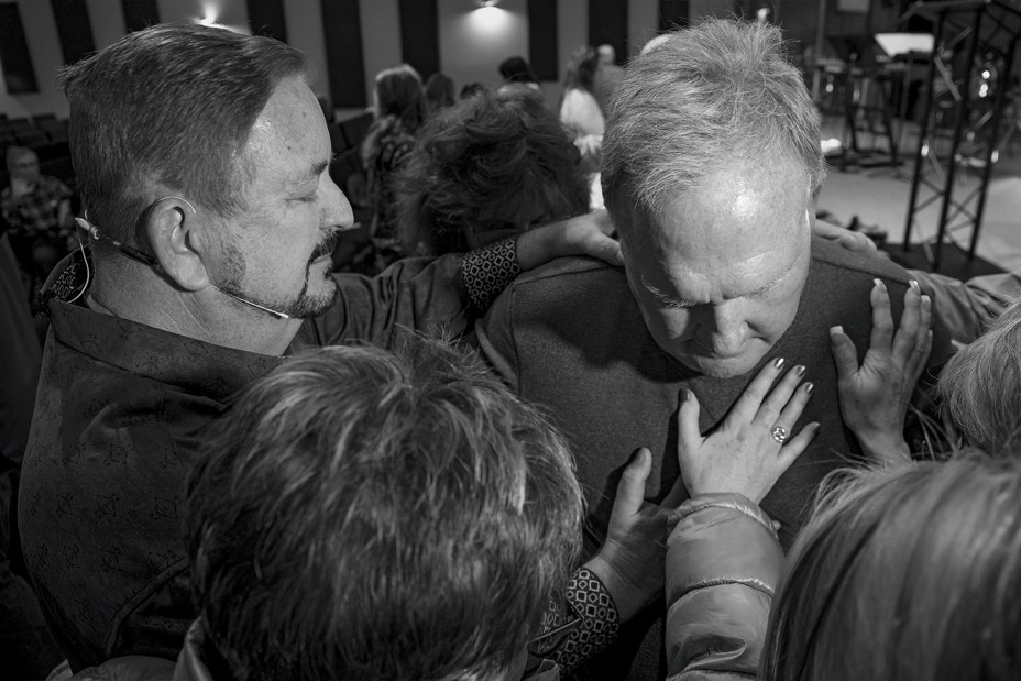 Photo of man with eyes closed and head down, with a number of other people placing hands on his body and praying