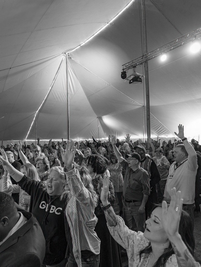 Photo of a crowd of people with eyes closed and hands and arms raised inside an enormous tent with stage lights