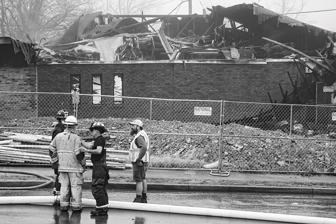 black and white photo of firefighters and person in safety vest talking in front of burned building with roof caved in behind a chain link fence