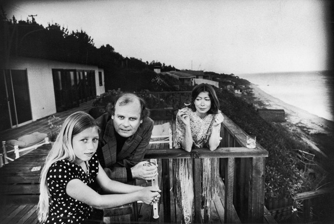 black and white family photo of Joan Didion on a deck at home, smoking and looking at her husband and Quintana Roo