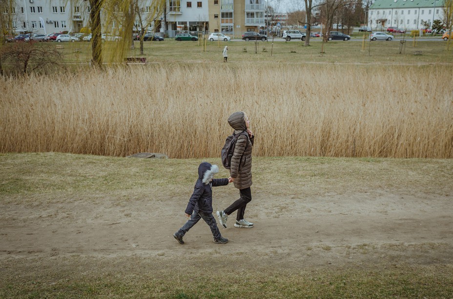 A woman and child wearing parkas and holding hands walking along a dirt path with grass and buildings behind