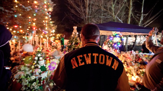 A man wearing a Newtown jacket visits a memorial.