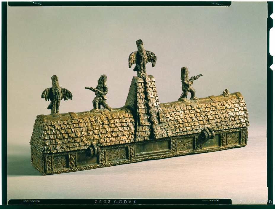 photo of metal sculpture of long rectangular building with steep roof and triangular tower in center, with figures of birds and warriors along roofline