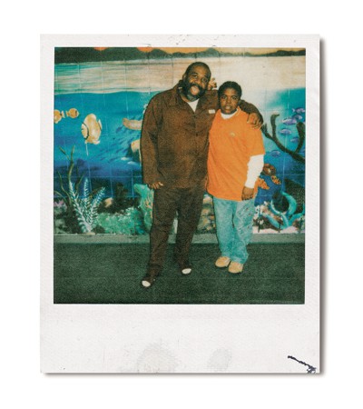 polaroid photo of man in heavy coat with arm around child in orange shirt and jeans standing in front of a mural of fish