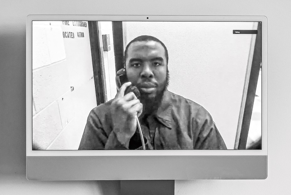 black-and-white photo of a seated, bearded man holding a phone receiver with a silver metal cord to his ear in a prison Zoom call 