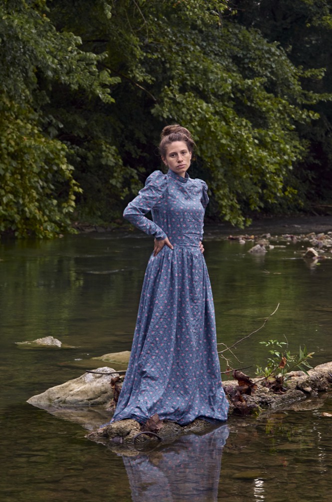 The photographer standing on rocks in a river wearing Victorian blue gown. 