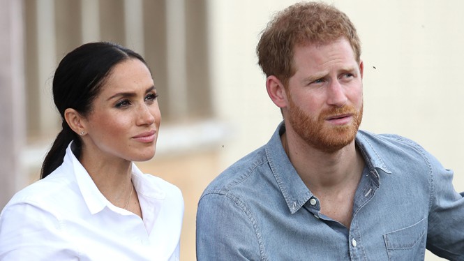 Meghan Markle, wearing a white button-up shirt with her hair slicked back into a low ponytail, and Prince Harry, wearing a chambray shirt, look off to the side of the camera.