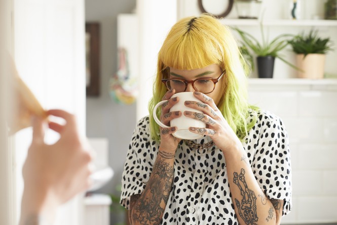 En ung kvinde med farvet hår og tatoveringer nipper til en kop kaffe.