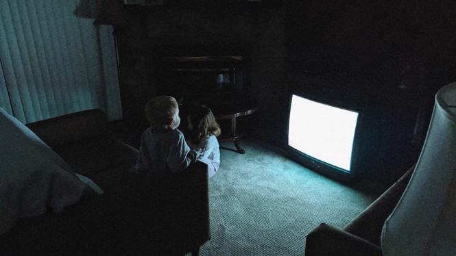 Two small children sit before a blank TV screen in a dark room.