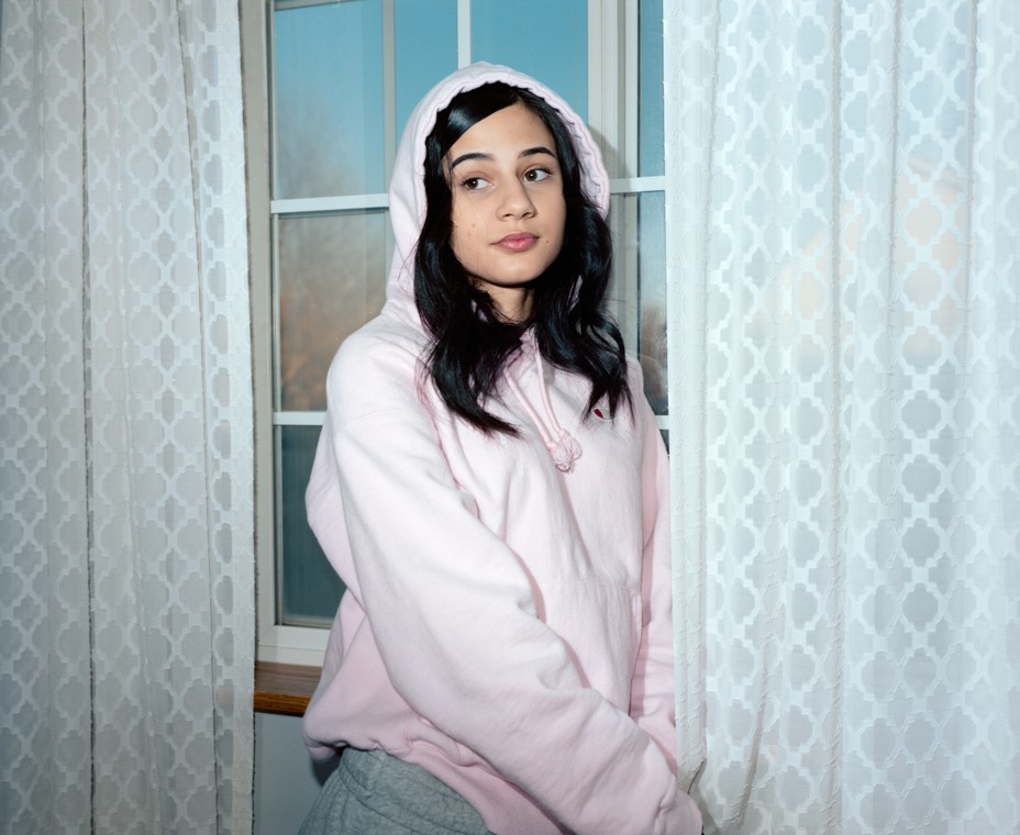 girl with dark wavy hair in light pink hoodie and sweatpants in front of window with long white curtains