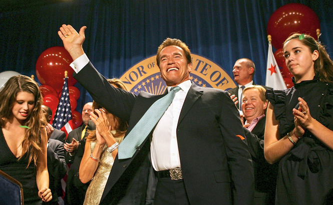 Photo of Schwarzenegger in suit raising arm, smiling and waving in front of state seal with flags and family applauding 