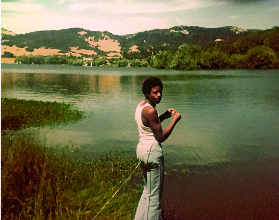 en mand ved en sø ser på kameraet og holder et fiskeri omgivet af bjerge omkring 1970'erne.
