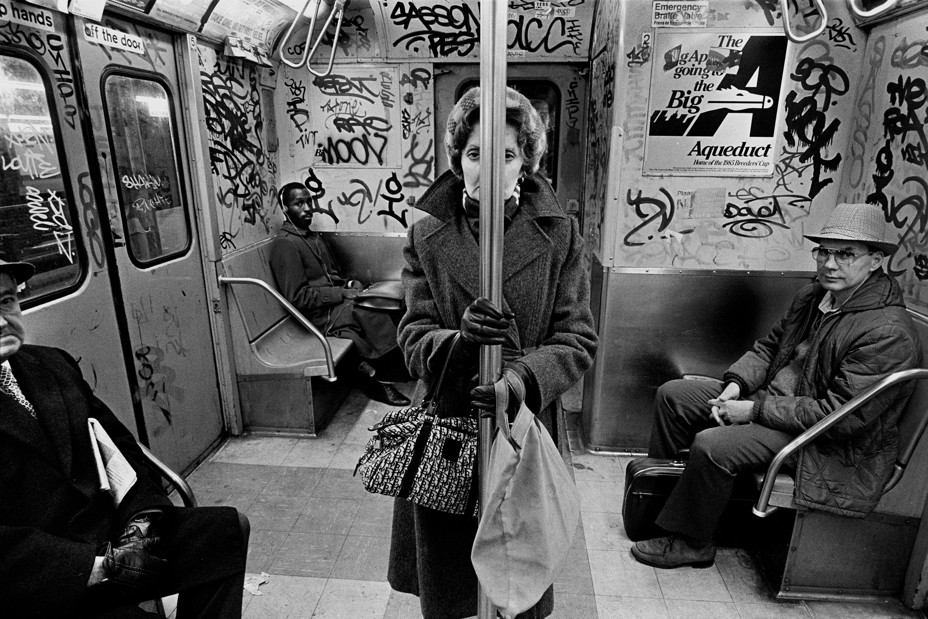 a woman on a graffitied subway car. she stares at the camera  with her face separated by the subway pole.