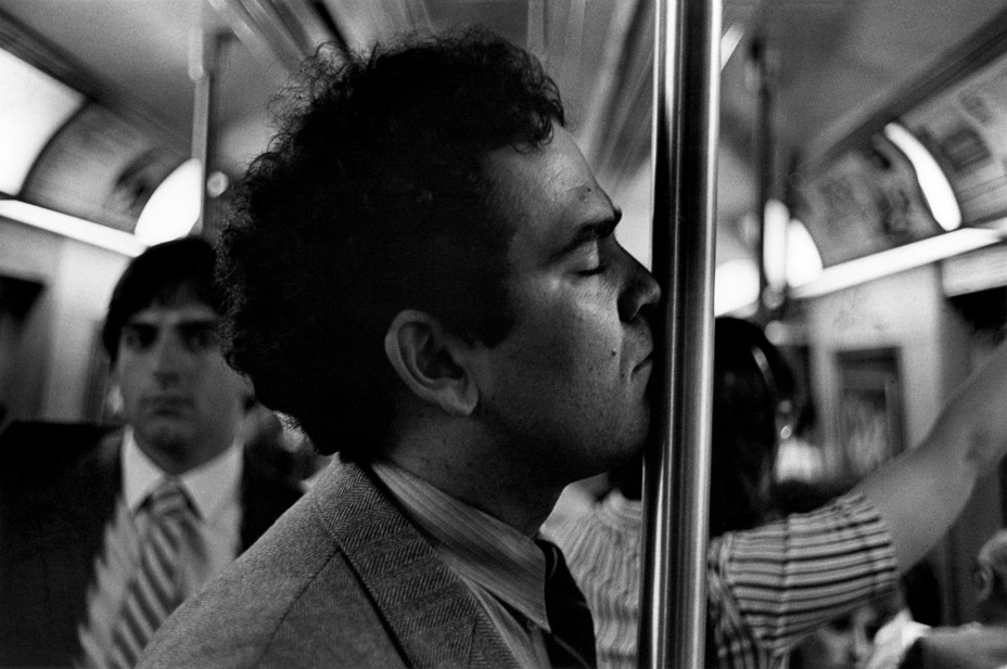 A man seen in profile leans his face into a subway pole with a man in the background looking at him.