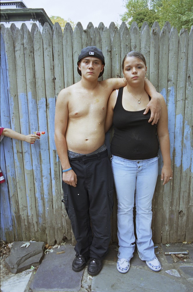 a young boy standing with his arms around a girl with a hand coming into frame with  a squirt gun