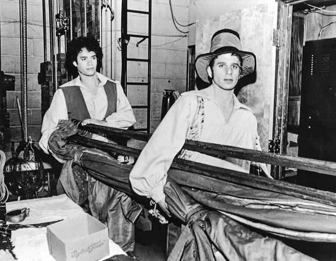 black-and-white photo of two costumed actors carrying scenery backstage