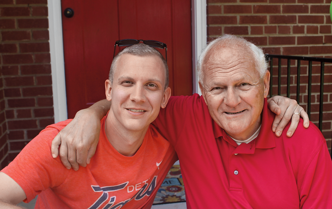 photo of younger and older man smiling with arms around each other in front of brick wall and door