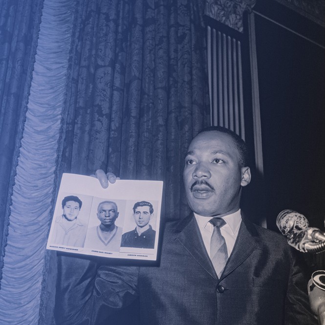 photo of MLK in suit and tie holding up picture of three slain civil rights activists