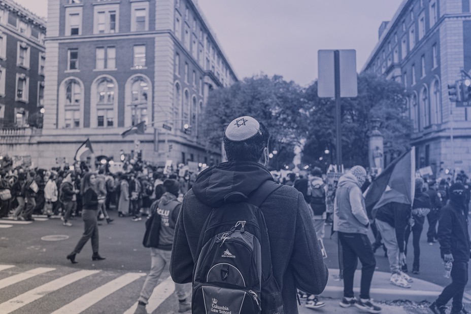 photo from back of student in kippah and backpack facing protest and people in street