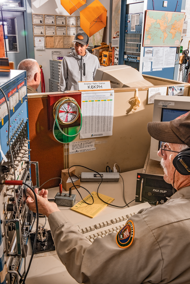 photo of uniformed man in headphones working in foreground with men in background talking