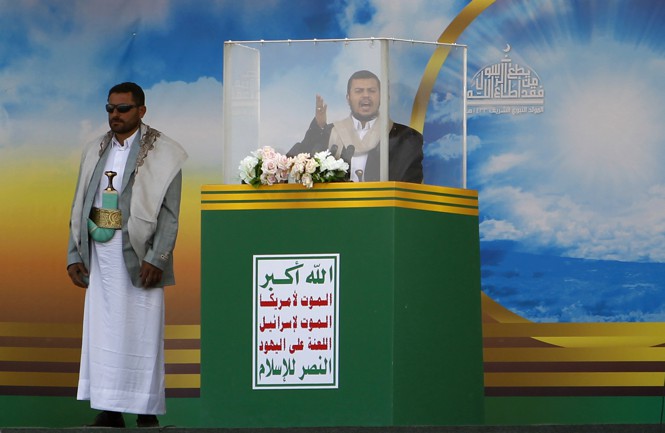 A man speaks in an enclosed podium standing in front of front of a blue sky wall paper