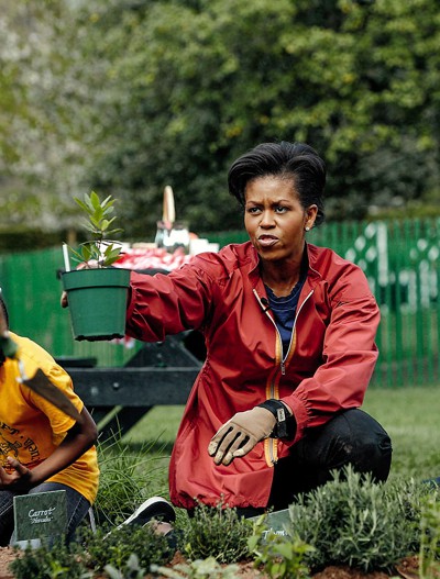 photo of Michelle Obama in red jacket kneeling and handing potted plant to volunteer in yellow shirt