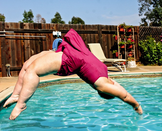 photo of woman in pink skirted swimsuit doing back dive into outdoor pool with fence in background