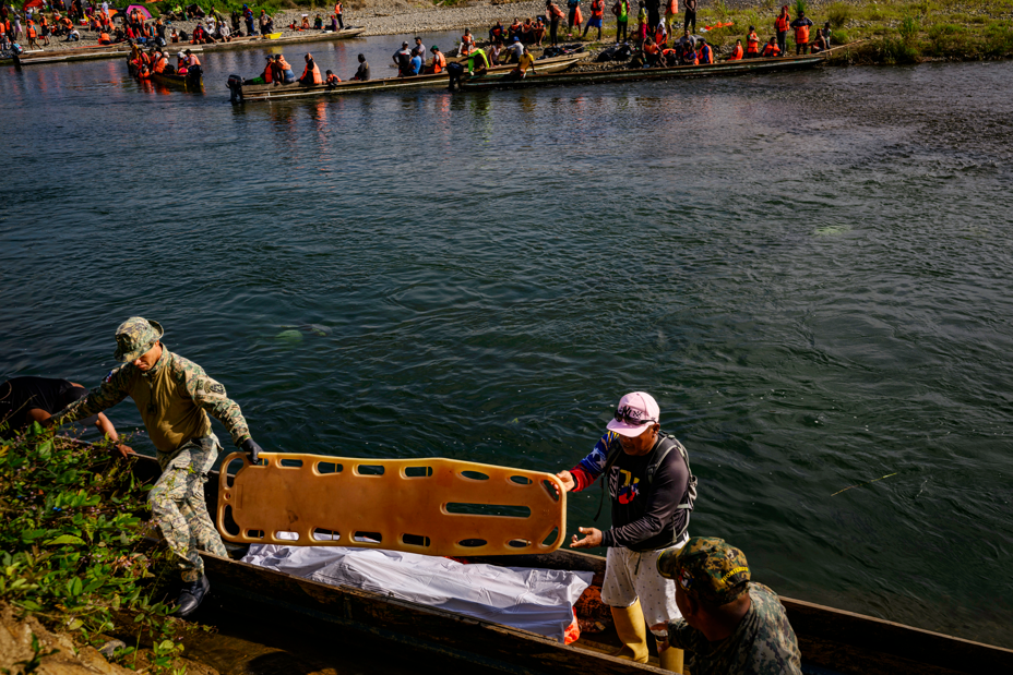 photo of two men holding stretcher next to very long thin boat with white body bag inside, with river and many other long boats full of people in background