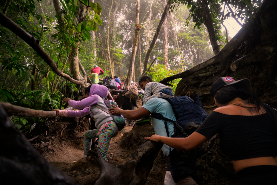 Foto de un hombre que lleva a un niño pequeño sobre los hombros ayudando a una niña pequeña a subir un sendero empinado y barroso en la selva, sujetándose de raíces largas, seguido por una mujer