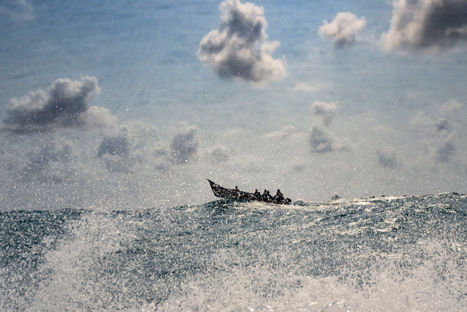photo of rough sea with spray, with prow of open boat full of people cresting a wave in distance