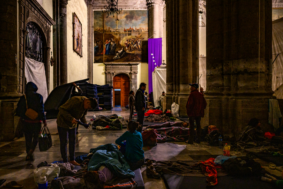 Foto del interior oscuro de una catedral, con columnas ornamentadas, tallas y pinturas, con muchas personas acostadas en mantas en el suelo