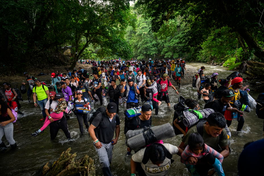photo of crowd of people wading through rocky stream, many carrying children and gear, with crowds more behind them surrounded by dense jungle