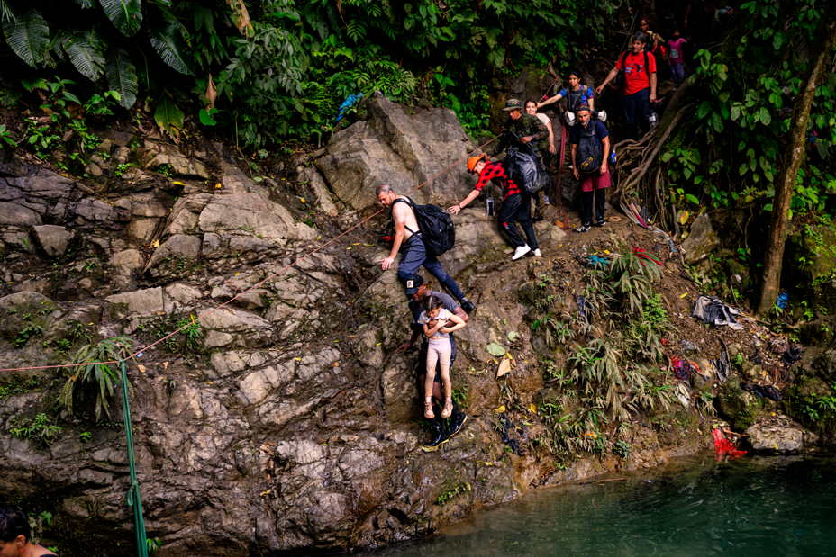 photo of man carrying girl with legs dangling along cracks in a very steep rock wall that drops away into water, with a line of people behind them waiting to also attempt to cross by holding on to a single horizontal rope