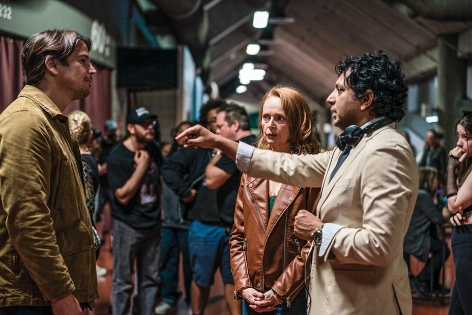 photo of Shyamalan in tan jacket gesturing toward Josh Hartnett as he listens, surrounded by cast and crew