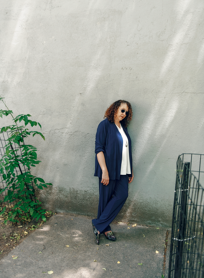 photo of woman with curly hair standing next to wall wearing dark glasses, white shirt, and blue pants and jacket