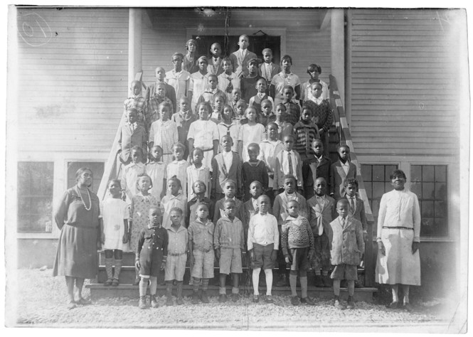 Dozens of students stand on the stairs of a school house with two teachers standing on the side.