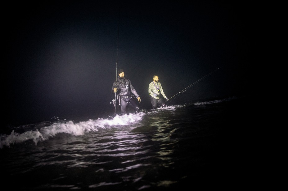 photo at night of two men in knee-deep surf holding fishing rods, lit by flash with black ocean and night sky around them