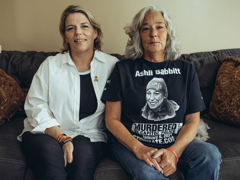 photo of two women sitting side-by-side on sofa with pillows; the woman on the right wears a t-shirt with picture of Ashli Babbitt and text "Murdered by Capitol Police"