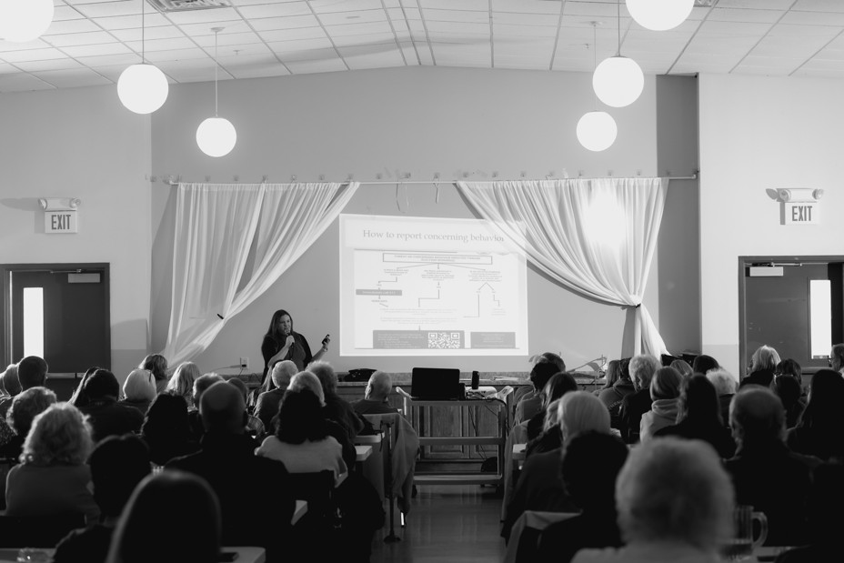 A black and white room of a person giving a presentation in front of a crowded room of people sitting