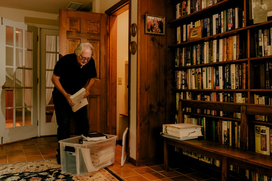 A white man hunched over holds a book, standing next to a bookshelf filled with books.