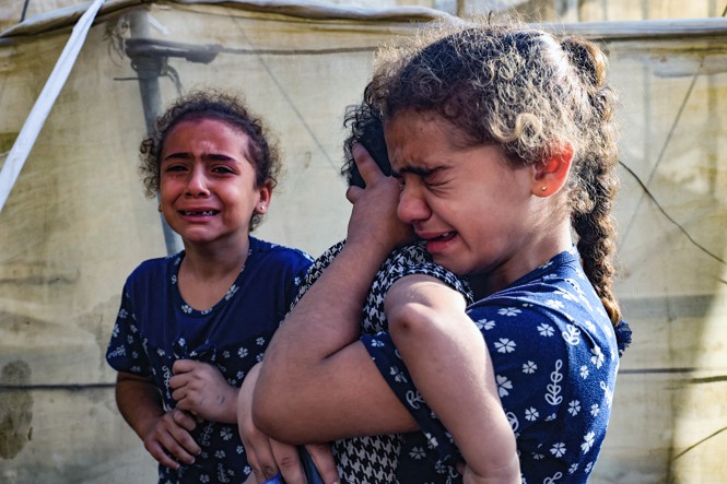 Three girls crying, all wearing blue shirts imprinted with white flowers, in front of a canvas wall.