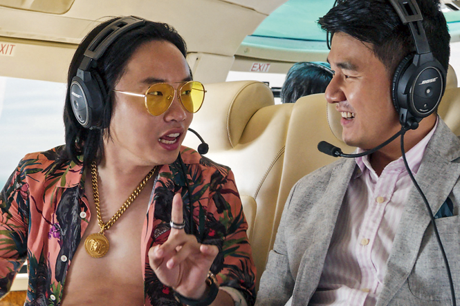 still from movie "Crazy Rich Asians" with Yang bare-chested in floral shirt with large necklace and yellow sunglasses talking to man in suit, both wearing headphones with mics