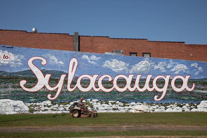 A man cuts grass in front of a mural of Sylacauga written on a wall.