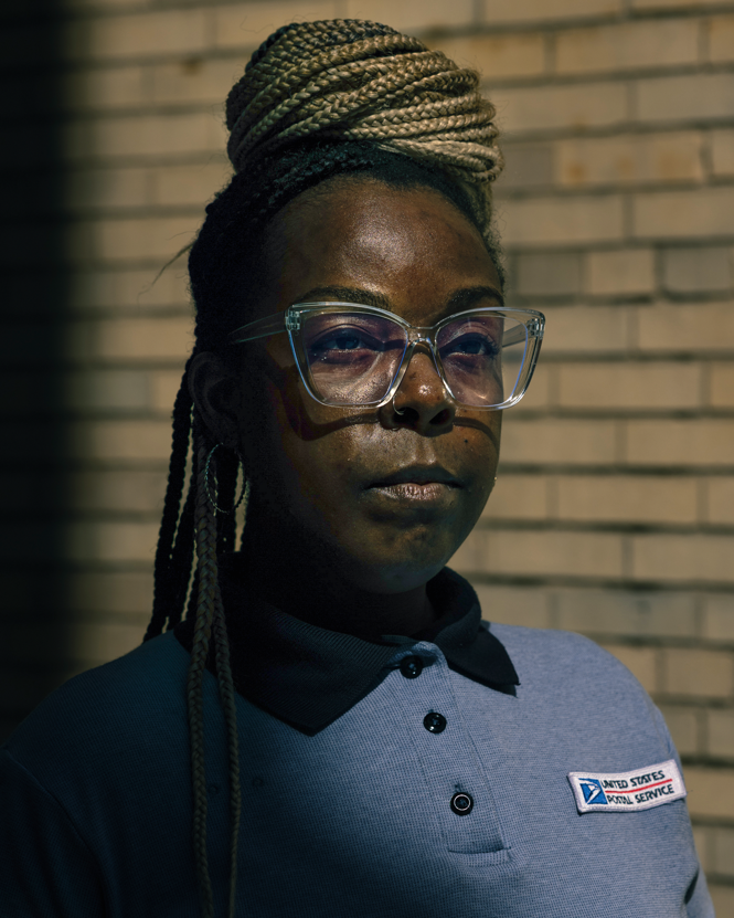 photo of woman with box braids in bun wearing large clear glasses and collared shirt with USPS logo