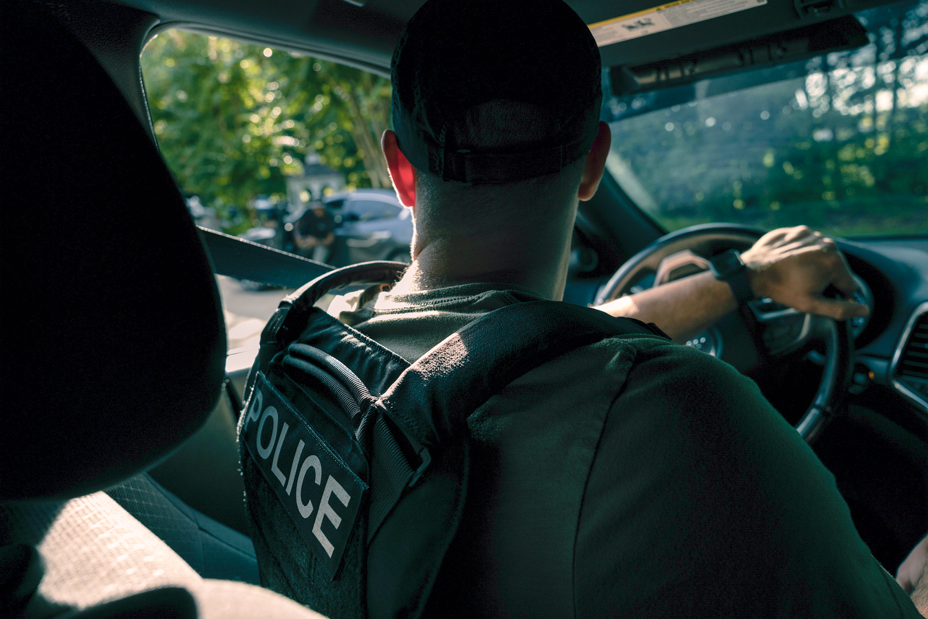 photo from back seat of man sitting in driver's seat of car with hand on steering wheel, wearing bulletproof vest that says 