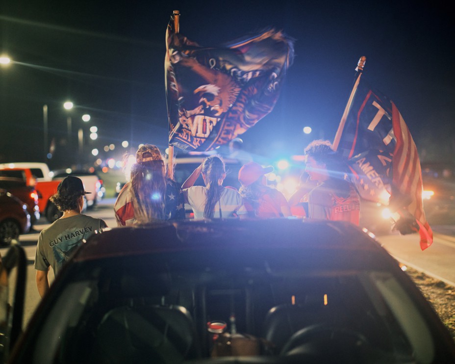 A group of five people are seen from behind sitting on a truck with flags and bright lights shining on them.