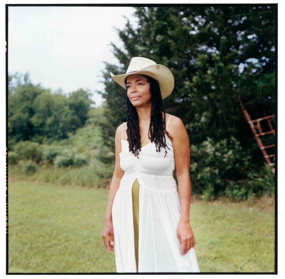 photo of Black woman with long hair wearing gauzy white sundress and straw summer cowboy hat standing in field with fence and trees behind