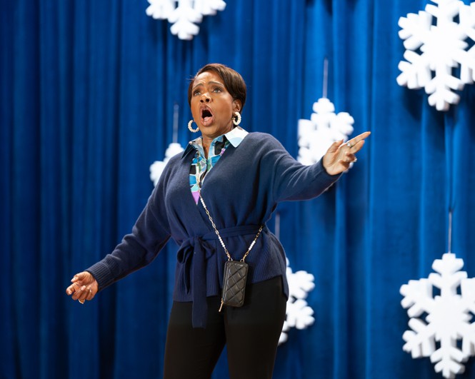 A woman sings with her arms extended wide on a stage with snowflake decorations and a blue curtain behind her