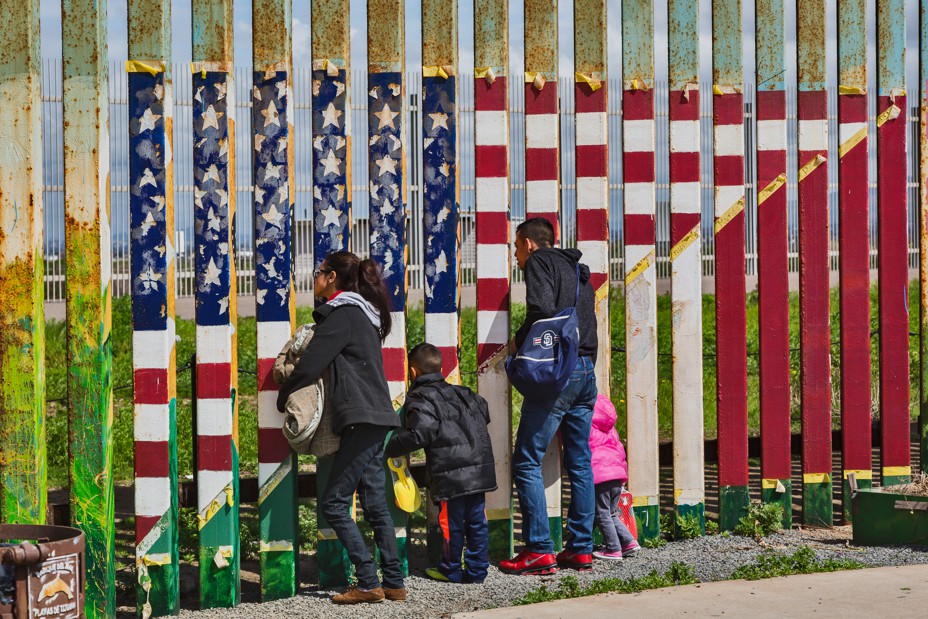 Two adults and two children look through the border fence that has an American flag painted on it.