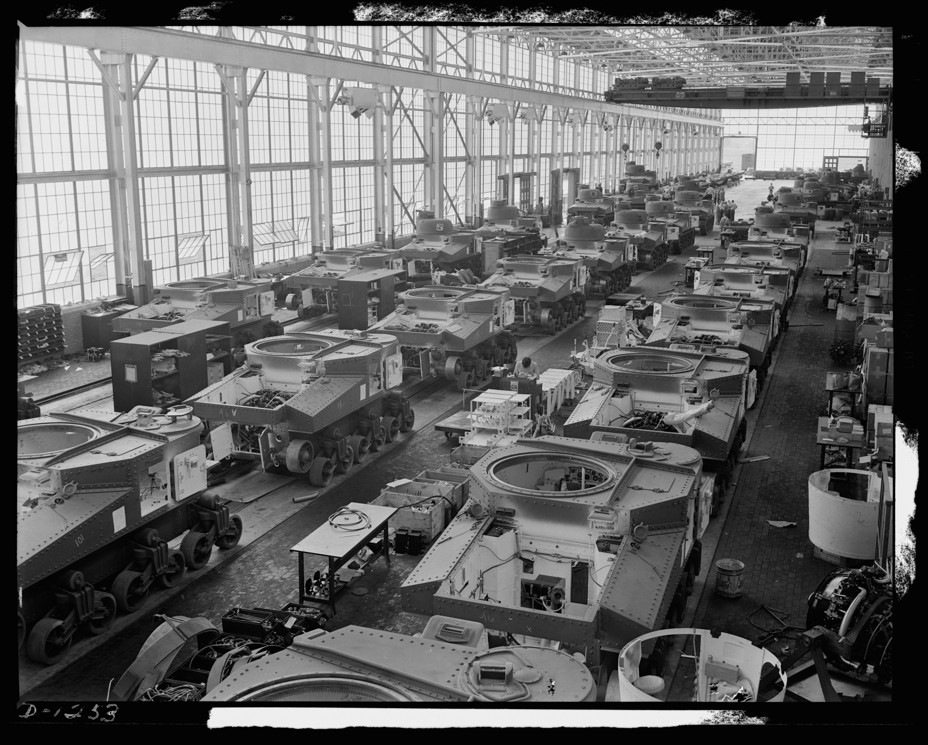 A black and white photo shows an assembly line of tanks lined up in a warehouse.